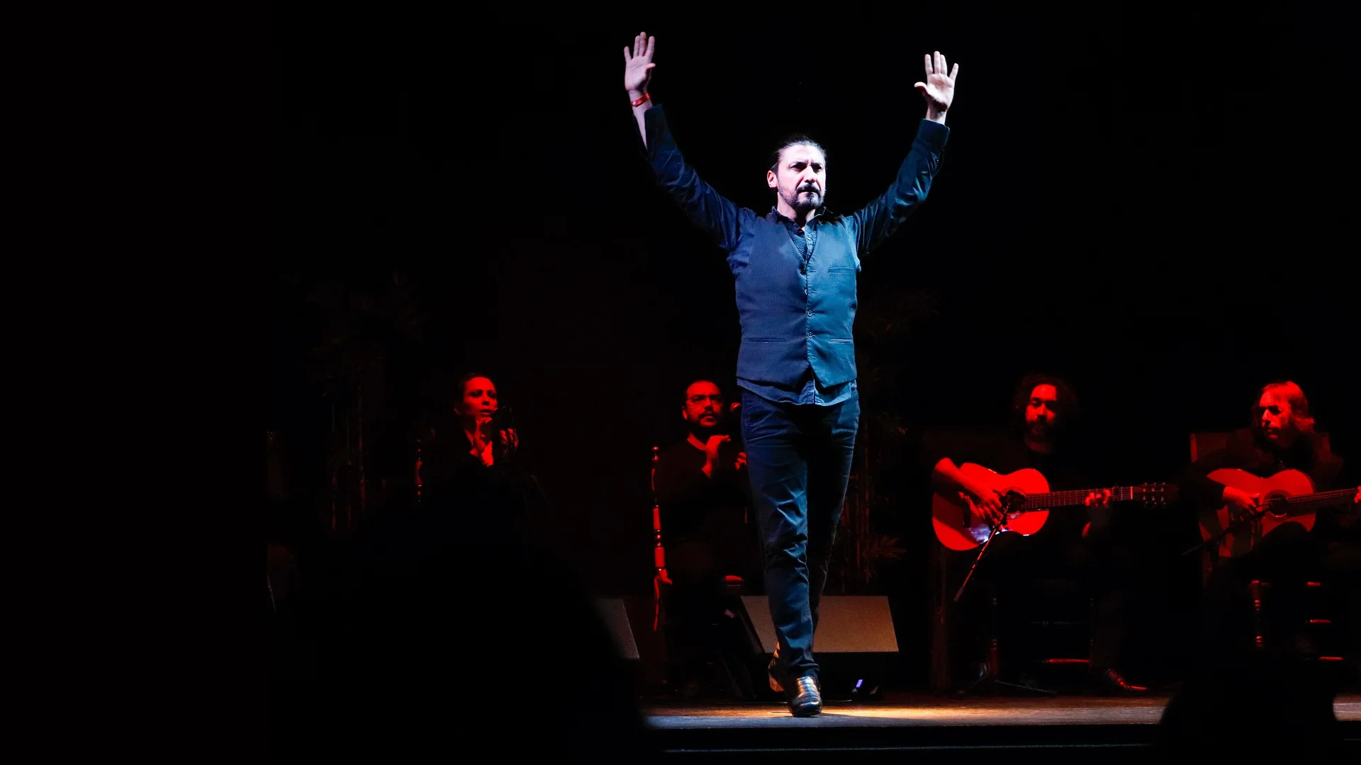 A flamenco dancer in a dark waistcoat stands with both arms raised on a dark stage, with a singer, a clapper and two guitarists seated in red light behind him