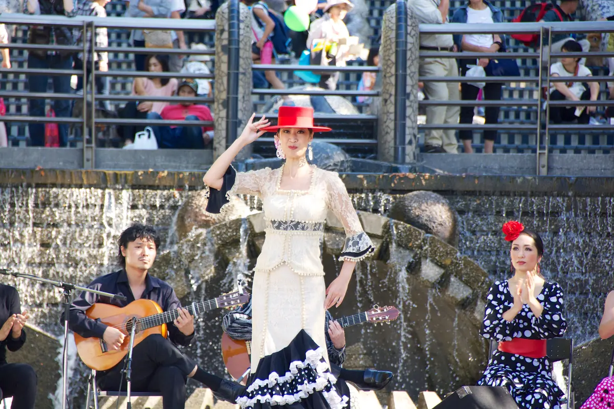 A flamenco dancer in a white lace dress and red hat between a guitarist and a woman clapping palmas